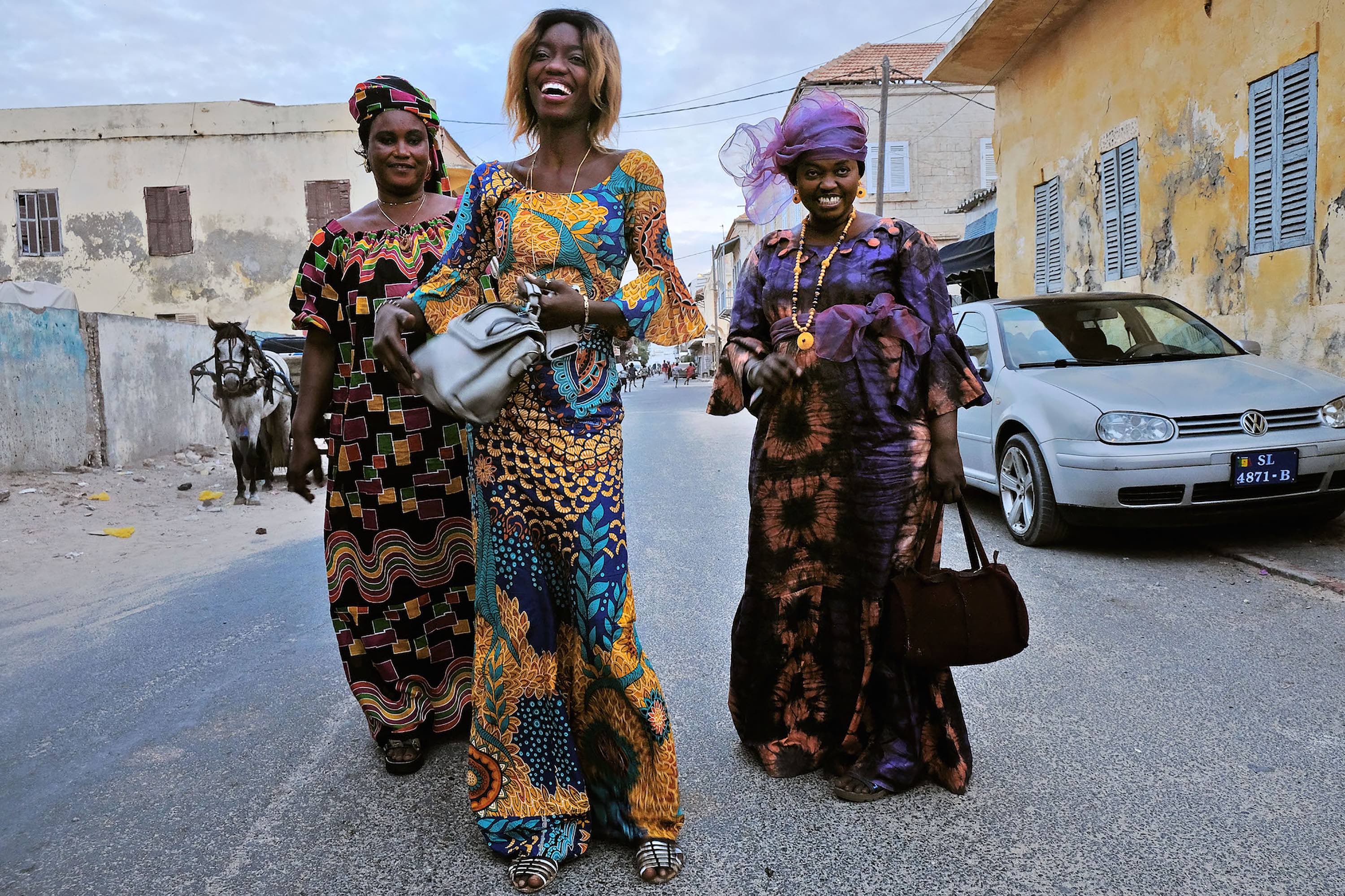 Three Senegalese women smiling wearing brightly colored stylish dress.